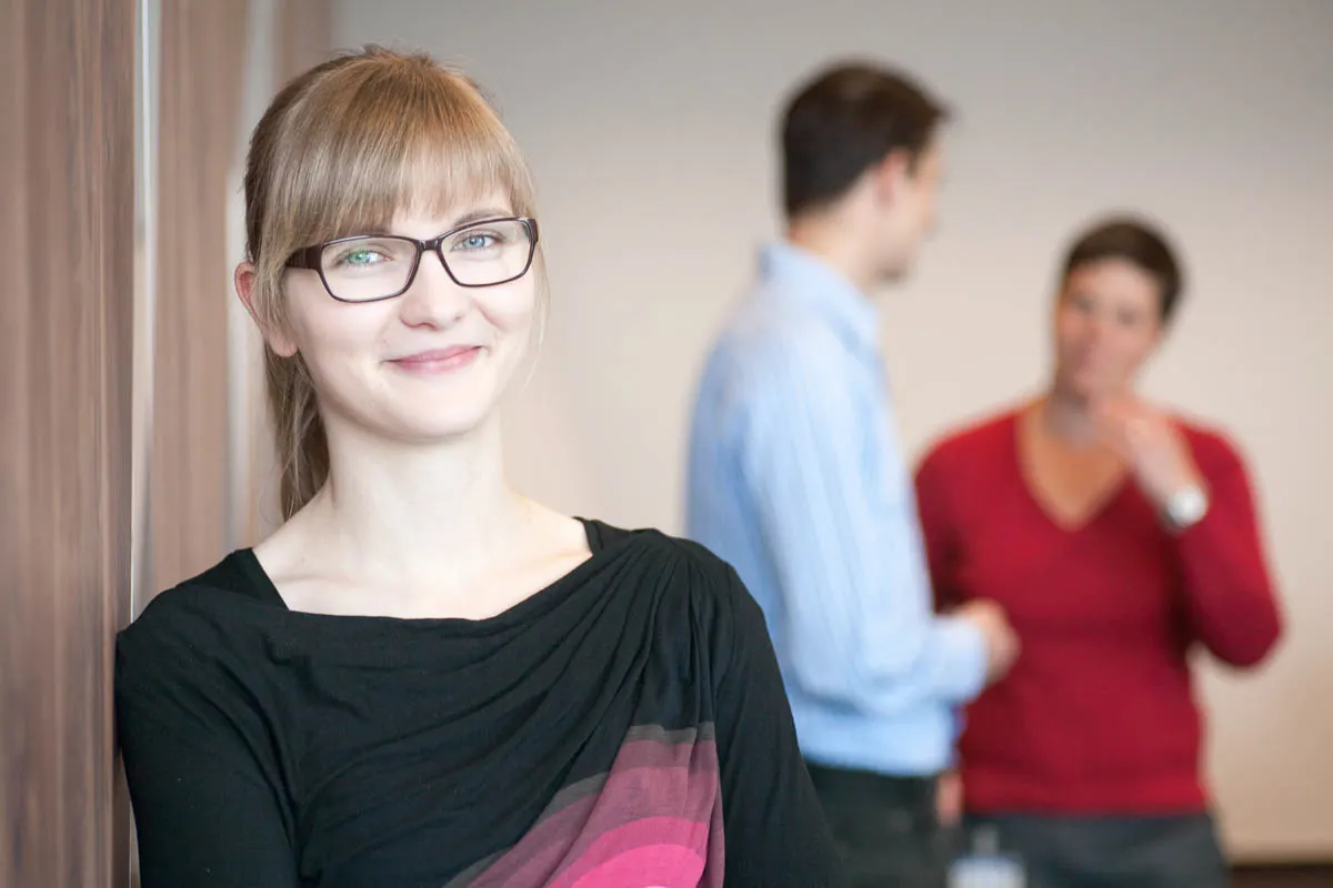 Mitarbeiterin mit Brille vor zwei unscharf dargestellten Kolleginnen und Kollegen in einem Büro.