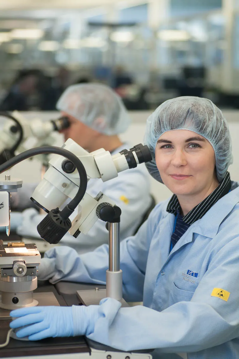 Mitarbeiterin in Schutzkleidung und Haarnetz arbeitet mit einem Mikroskop im technischen Arbeitsumfeld