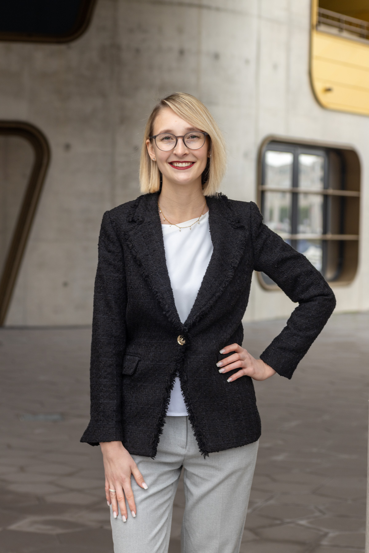 Businessportrait: Blonde Frau mit Brille, schwarzem Blazer und heller Hose vor einer modernen Gebäudefassade.
